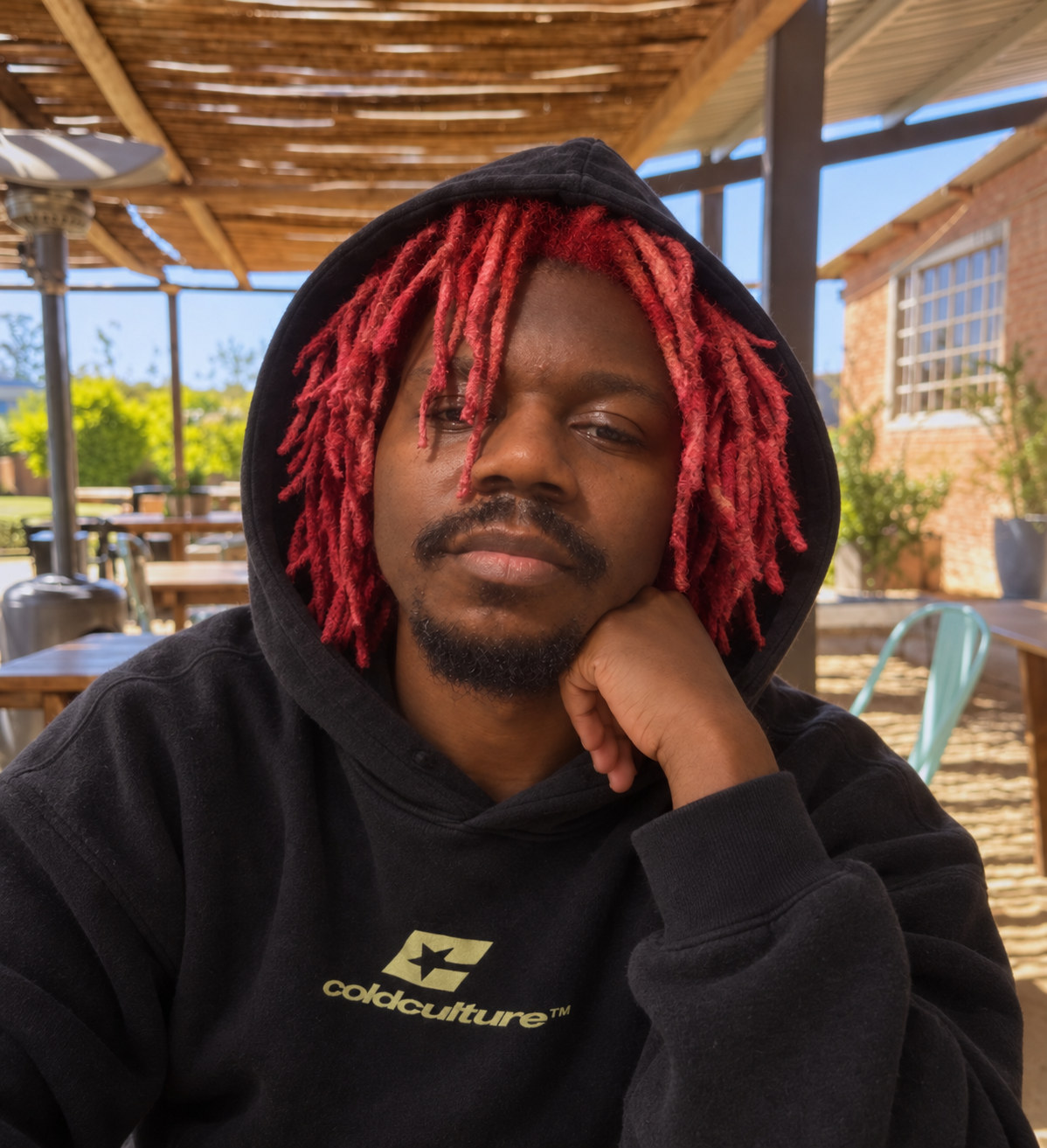 Prince Desire in a black hoodie with red locs, hand on chin, outdoors.
